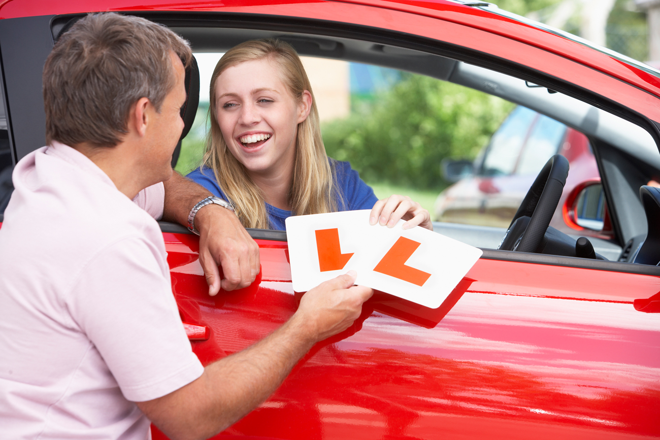 A young girl in the driver seat of a car with her dad holding learner plates